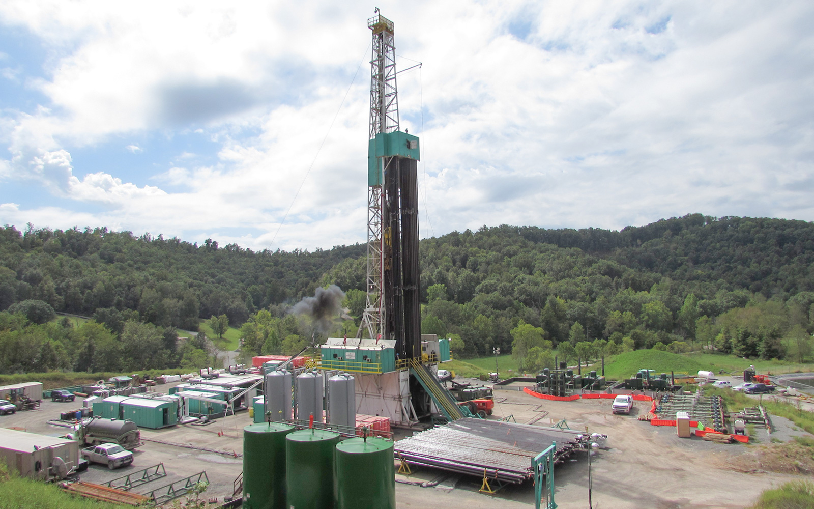 A drilling rig set up in a cleared dirt lot with trucks and trailers around, and trees in the background