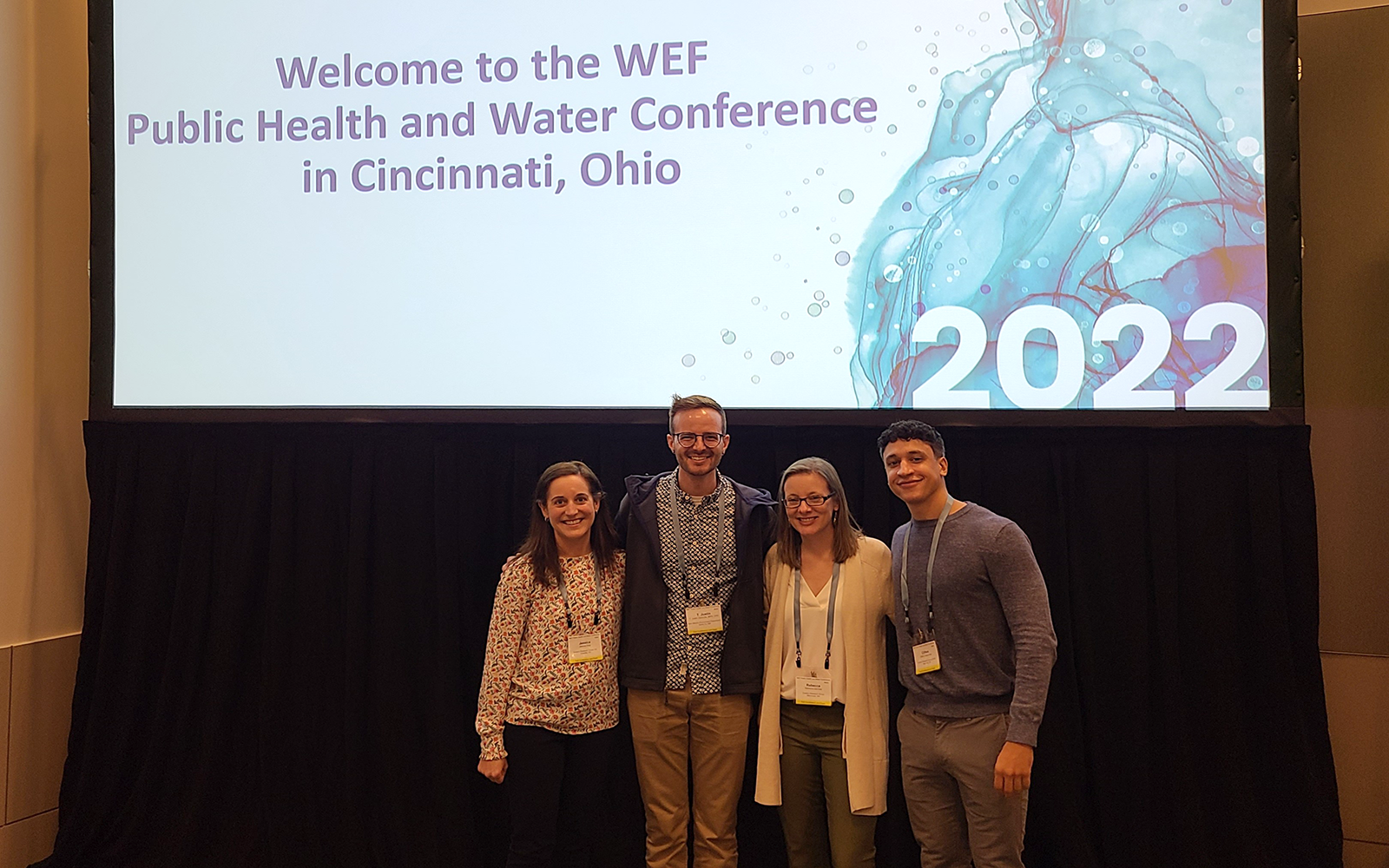 Four people (two men and two women) pose in front of a sign that reads Welcome to the WEF Public Health and Water Conference in Cincinnati, Ohio.