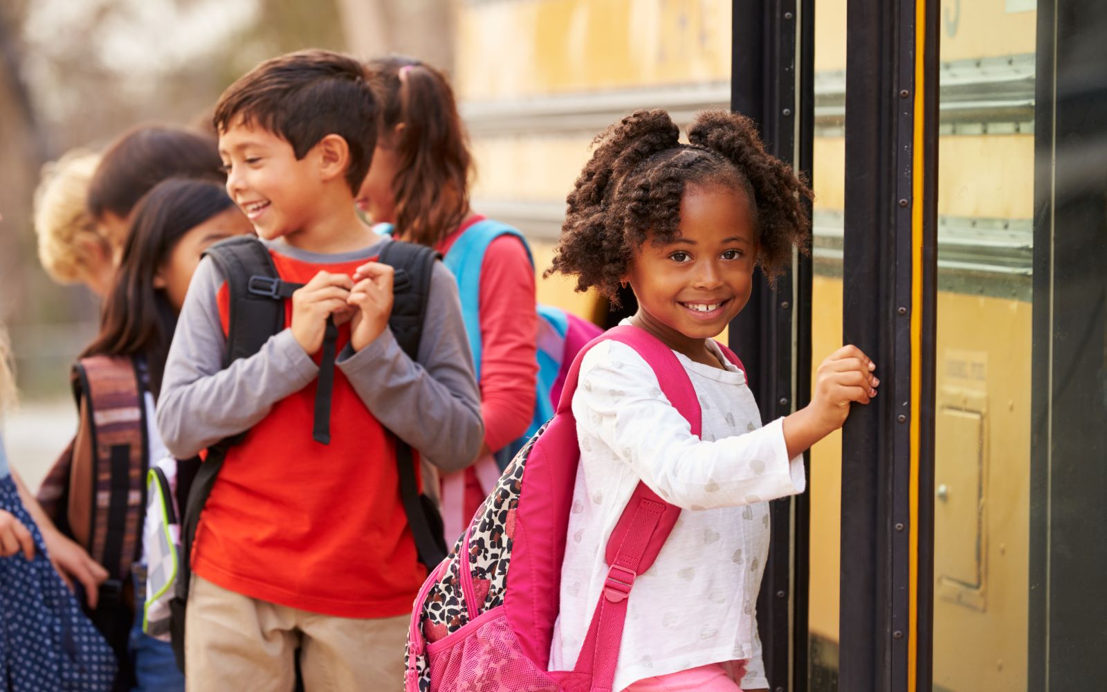 Children lining up in front of a school bus, smiling and wearing backpacks