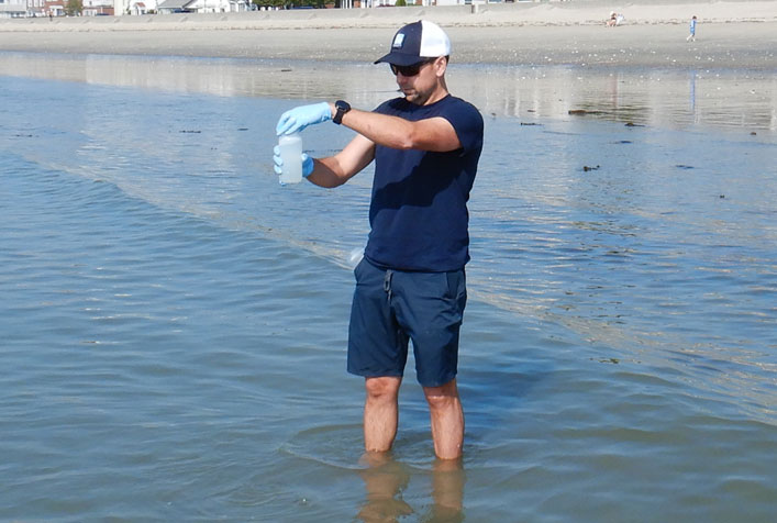 Photo of person standing ankle deep in ocean shore wearing latex glove collecting a water sample in a small container