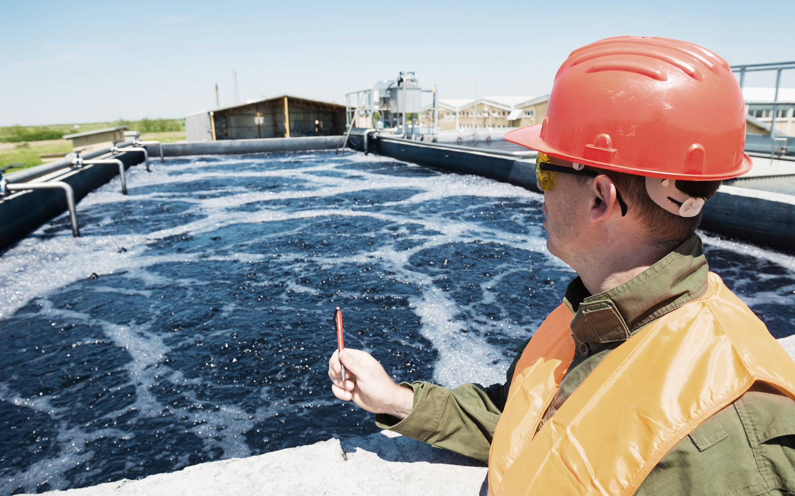 Man in hard hat and safety glasses looking at water in a wastewater treatment tank.