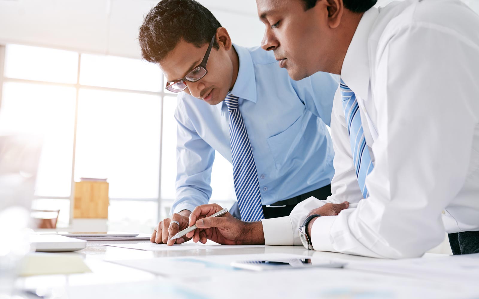 Two men standing side by side over an office table carefully examine the contents of a document open on the table.