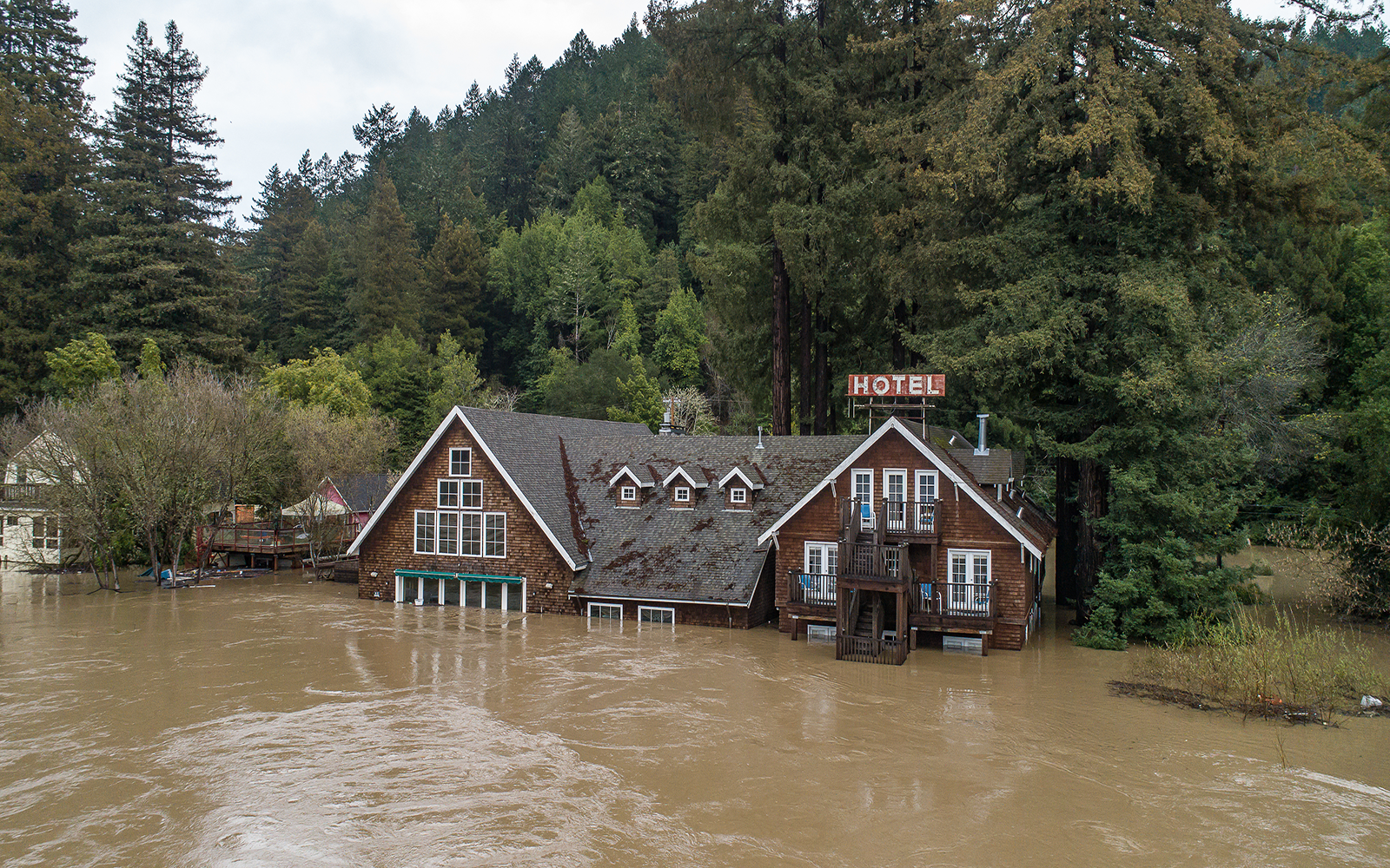 A hotel building is half-concealed by brown floodwater in a wooded area.