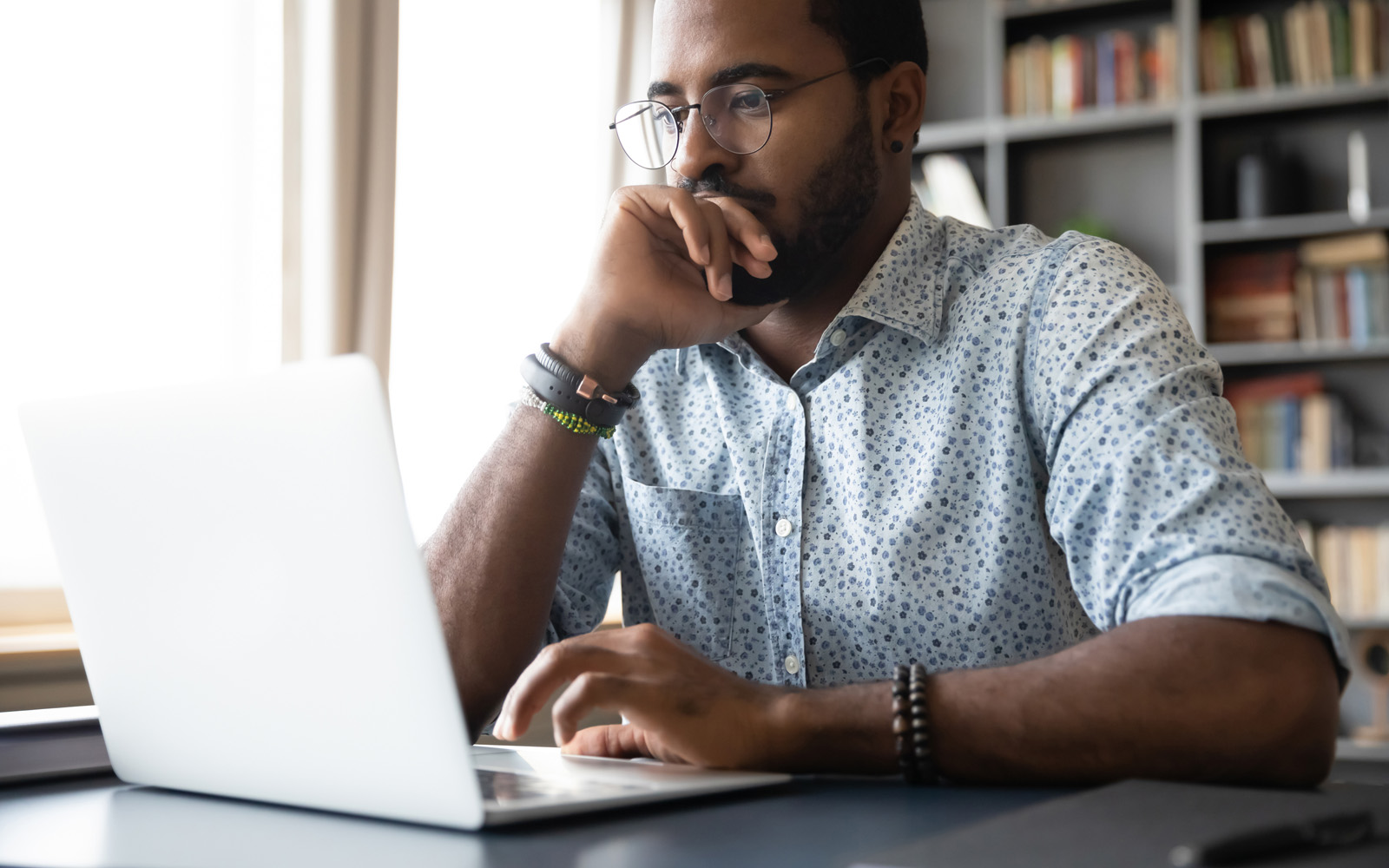 Man with beard and wire-rimmed glasses sits at laptop, thinking.