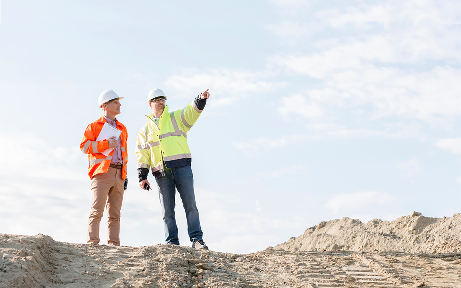 Two construction workers in orange and yellow vests and hard hats survey the nearby land