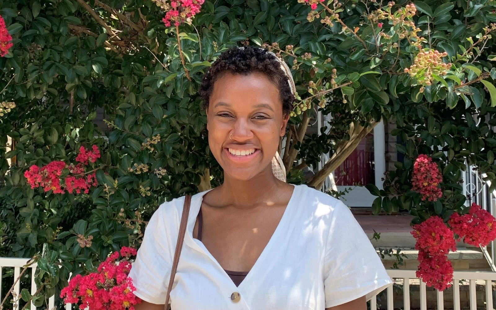 Deja, a Black woman in a white blouse, stands in front of a pink crepe myrtle tree