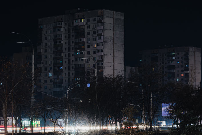 Photo of a tall city building at night during a blackout with only the street traffic as illumination seen through the trees 