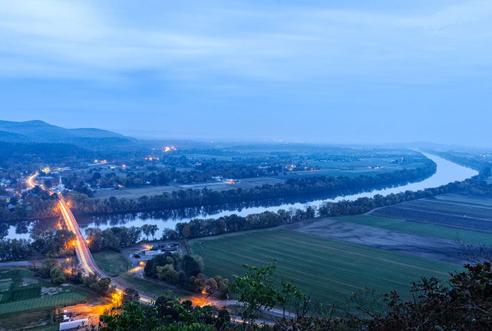 Photo of a river viewed from a distance with a lighted roadway bridge passing over it.