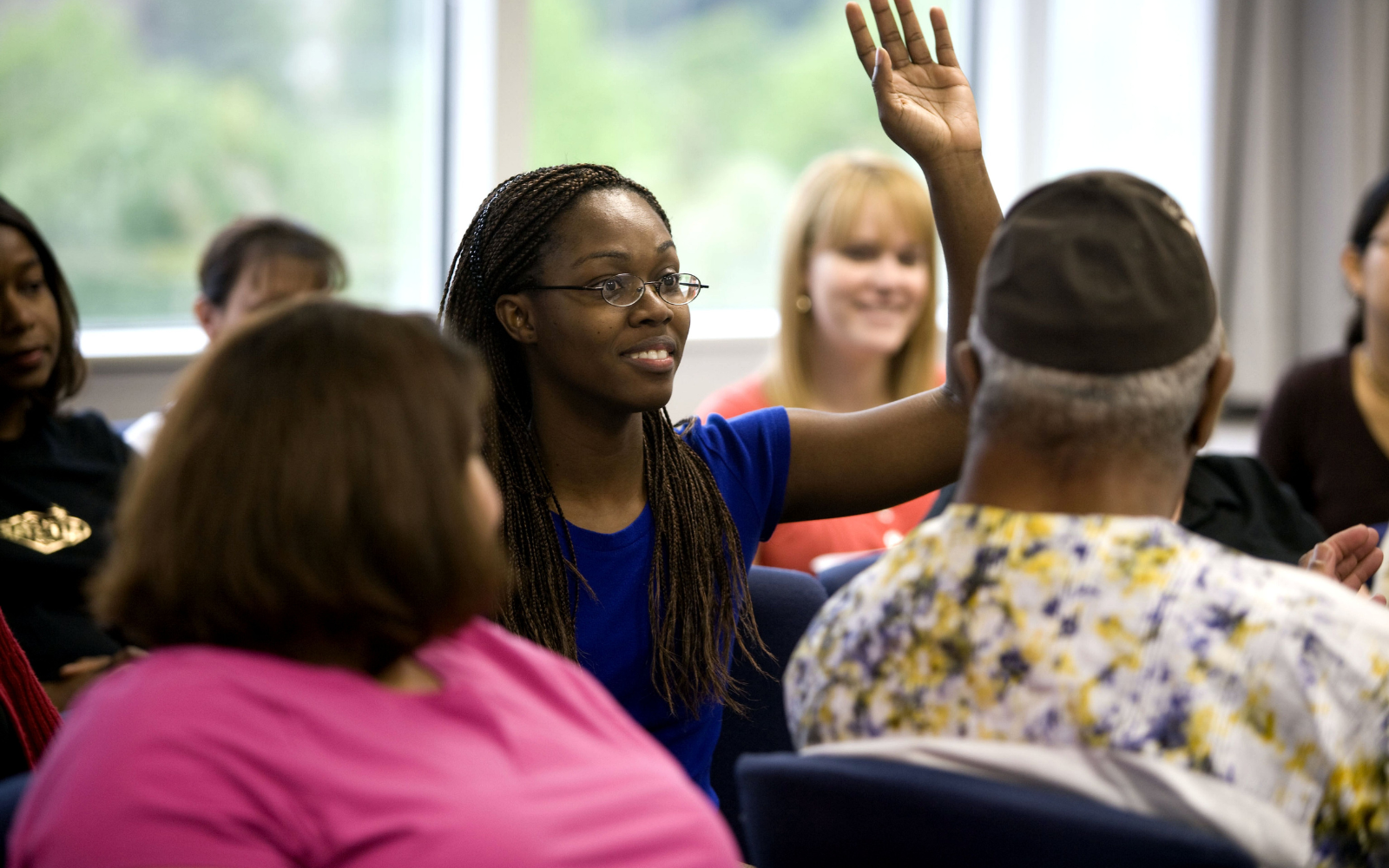 A group of people sit in a meeting room. In the middle, a Black woman raises her hand.