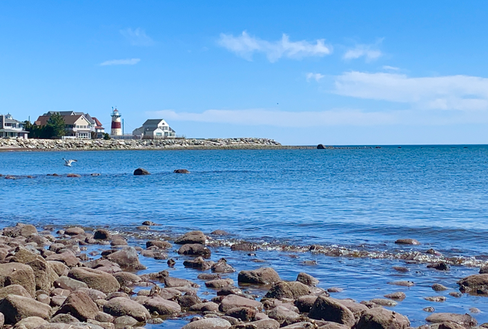 Photo of the Long Island Sound. image contains rocky shore, the water, a lighthouse and houses in distance