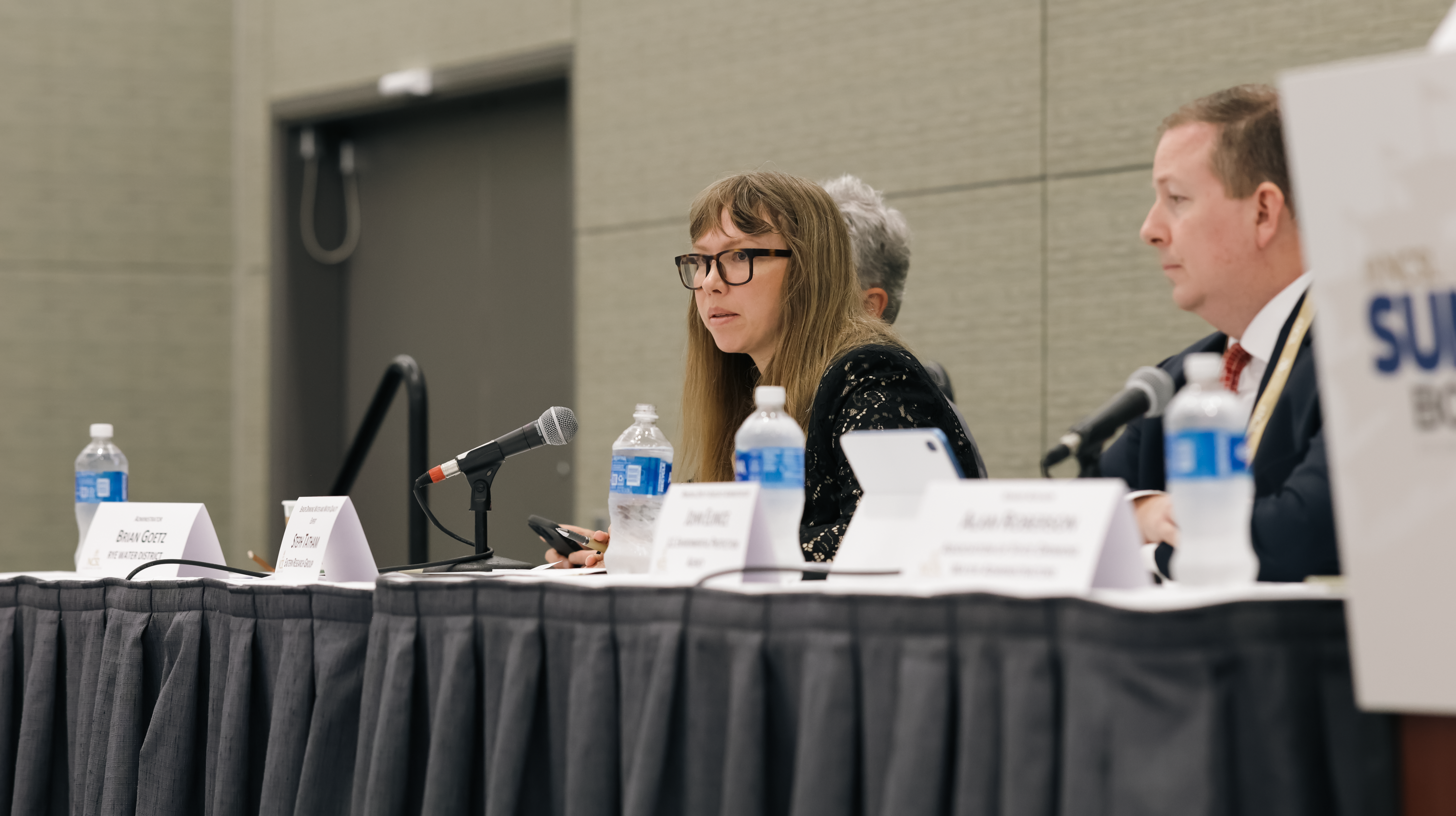 Woman with long hair and glasses leans into a mic at a table with a black tablecloth