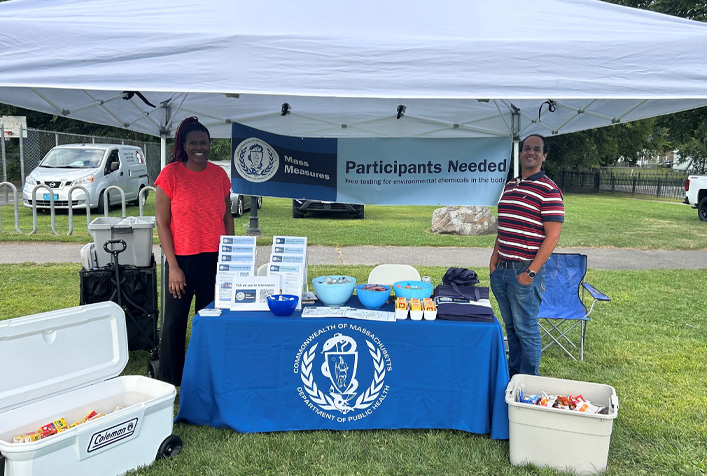 Image of two people standing under a white tent with a table of items and a Commonwealth of Massachusetts logo on a banner that reads Participants Needed