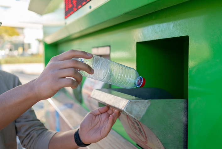 arms of a person in the process of depositing an empty plastic water bottle into a large public recycling waste container
