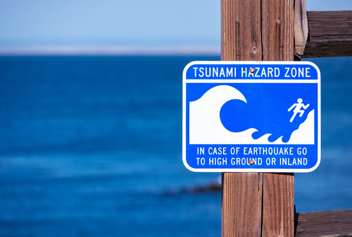 Photo of a warning sign by the shore that states: "Tsunami Hazard Zone, in case of earthquake go to high ground or inland."